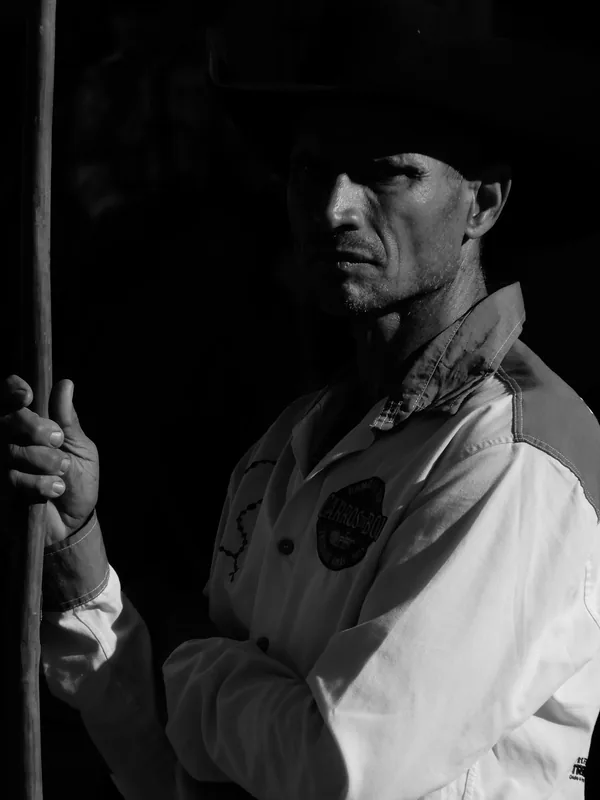 Fotografia em preto e branco, no formato vertical, mostrando um homem enquadrado do peito para cima, em um ambiente predominantemente escuro.
